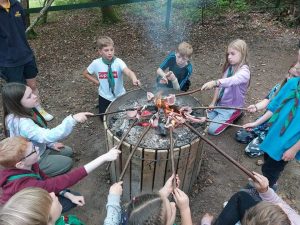 Young people in a circle cooking bacon on a stick over a bin fire